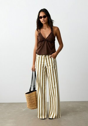 Femme portant un haut sans manches marron foncé, un pantalon large rayé, des lunettes de soleil, et tenant un sac fourre-tout tissé sur un fond uni.