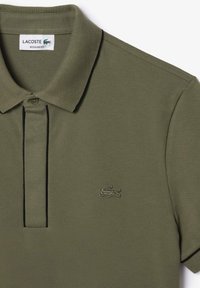 Polo de color verde oliva con tejido texturizado, cuello acanalado, tapeta de tres botones y un ribete negro. Logo de Lacoste bordado en el pecho.