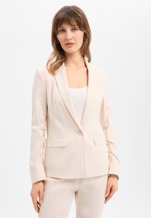 Vrouw met schouderlang haar die een lichtbeige op maat gemaakte blazer en bijpassende broek draagt tegen een effen witte achtergrond.