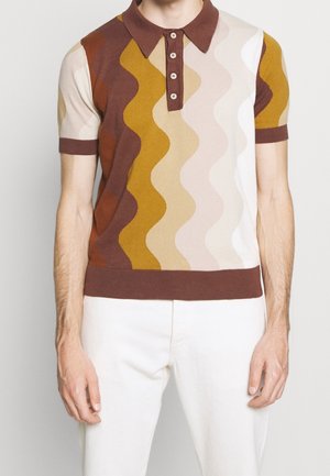 Mann trägt braunes Poloshirt mit kurzen Ärmeln und wellenförmigen Streifen in Braun, Senfgelb, Beige und Weiß, kombiniert mit weißer Hose.