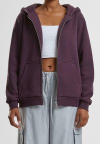 Personne portant un sweat à capuche zippé violet foncé par-dessus un débardeur court blanc et un pantalon cargo gris clair avec une taille à cordon.