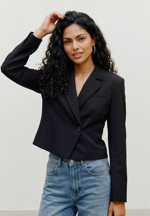 Jeune femme aux cheveux noirs bouclés, portant des boucles d'oreilles anneaux, un blazer noir court et un jean bleu, souriante avec une main touchant ses cheveux.