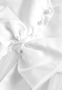 Bílá saténová látka stažená do velké mašle, ozdobená třemi perlovými knoflíky na hladkém materiálu.