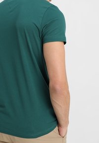 Turkoosi puuvillainen t-paita lyhyillä hihoilla; siinä on pyöreä kaula-aukko ja sileä pinta. Takapuolelta ei näy näkyviä logoja tai kuvioita.