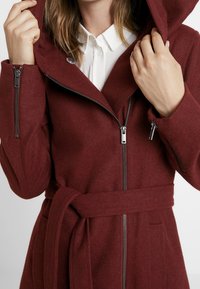 Bordový vlněný kabát s páskem v pase, kapucí a zipovými předními kapsami. Obsahuje zipové zapínání a měkkou, teplou texturu.