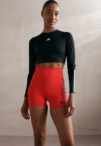 Crni dugih rukava crop top s bijelim logotipom, uparen s crvenim kratkim hlačama visokog struka koje imaju sive bočne pruge i isti logotip sprijeda.