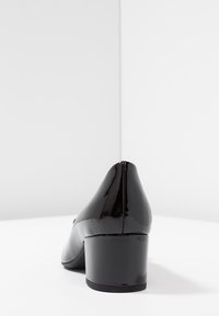 Svart lackläder lågt blockklackssko sedd bakifrån på en vit yta mot en vit hörnvall.