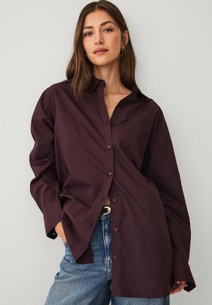 Kvinde iført en løs mørk lilla skjorte med knapper og blå jeans, stående mod en almindelig lys væg med et afslappet udtryk.