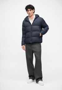Jachetă puf navy cu glugă, cu un fermoar frontal, design cusut și accent cu logo. Asortată cu pantaloni gri și adidași albi.