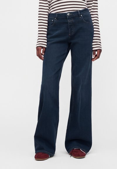 Personne portant un jean bleu foncé à jambes larges, une chemise rayée blanche et marron à manches longues et des chaussures rouges, debout devant un fond uni.