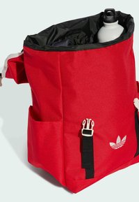 Zaino in tessuto rosso con interno nero, dotato di tasche esterne, hardware bianco e un logo stampato sulla parte anteriore.