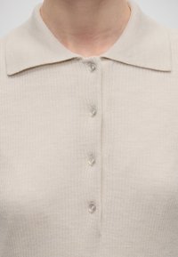 Pull en tricot beige avec col large et quatre boutons translucides rapprochés sur le devant, porté par une personne visible du cou à la poitrine.