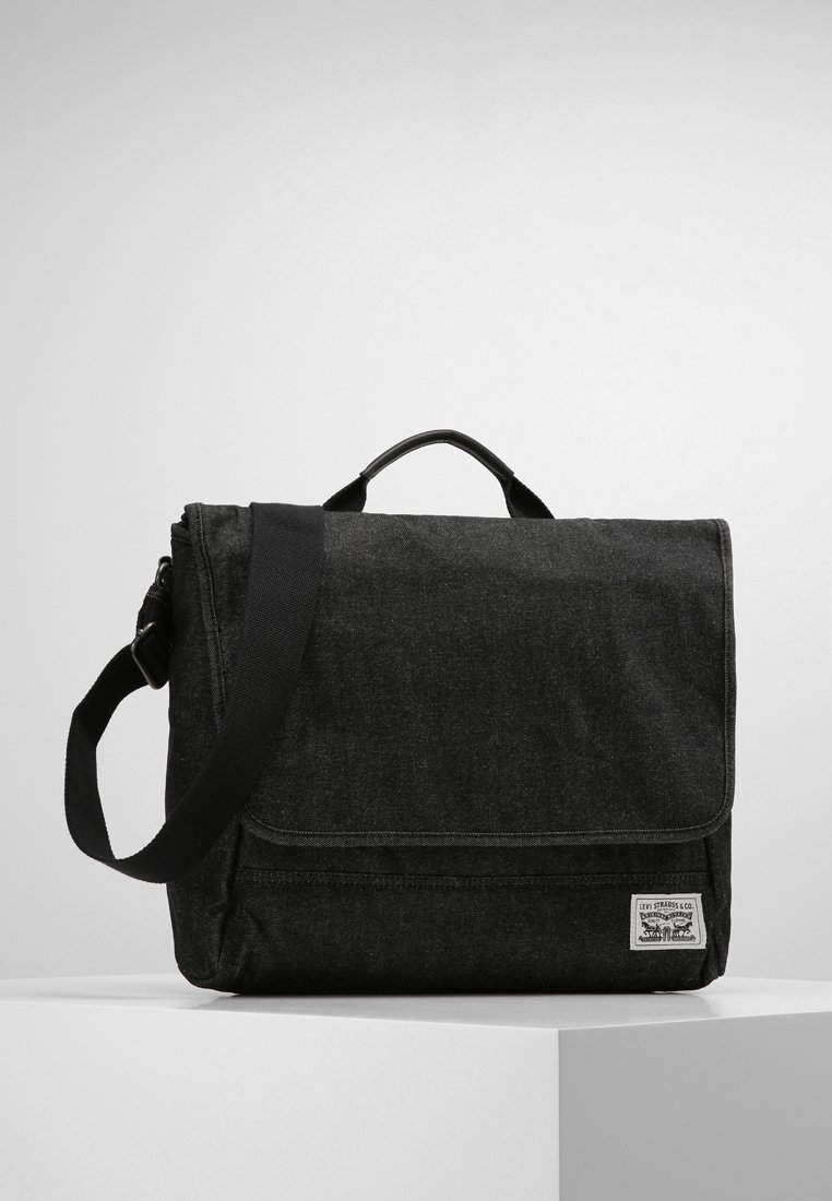 Bolsa de mensajero negra de lona con cierre de solapa, correa ajustable, asa superior y parche con logo en la parte frontal. Superficie plana y texturizada con detalles de costura.