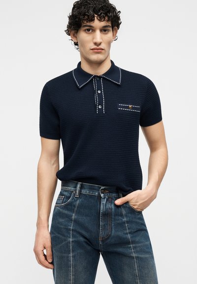 Bărbat purtând un polo tricotat cu mâneci scurte, de culoare bleumarin închis, cu detalii cusute albe și blugi bleu închis, stând cu o mână în buzunar.
