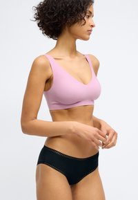 Lichtroze bralette met een diepe V-halslijn; gecombineerd met zwarte slipjes met kant langs de tailleband. Glad materiaal overal.