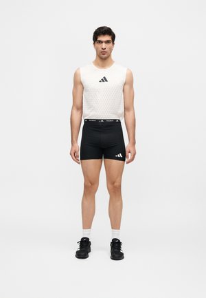 Muž stojící čelem vpřed, oblečený v bílém tílku Adidas, černých kompresních šortkách Adidas, bílých ponožkách a černých teniskách.