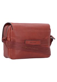 Borsa a tracolla in pelle arancione bruciato, con un patch logo frontale, tracolla regolabile con nome del marchio e chiusura superiore con zip.