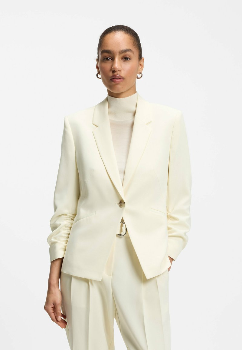 Femme portant un blazer crème sur mesure et un pantalon assorti, les mains dans les poches, debout devant un fond blanc uni.