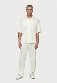 Camisa de botones de manga corta en color crema con dos bolsillos en el pecho, combinada con pantalones de tiro elástico a juego y zapatillas blancas. Textura de tela suave.