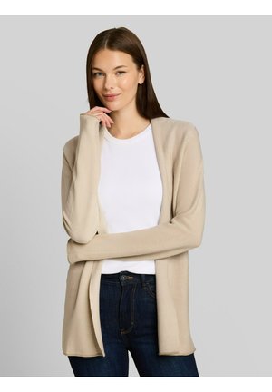 Junge Frau mit glatten braunen Haaren, die eine beige offene Strickjacke, weißes Top und dunkelblaue Jeans trägt, posiert mit einer Hand in der Nähe ihres Gesichts.