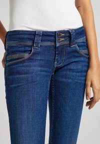 Blue denim jeans with a fitted design, featuring a high waist, two-button closure, and contrasting stitching. Pockets visible on the front.
