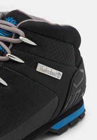 Černé turistické boty s šedými tkaničkami, kovovými detaily a modrým akcentem. Vyznačují se texturovaným svrškem a robustním designem podrážky. Logo Timberland je přítomno.