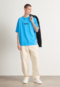 Svetlá modrá voľná t-shirt s čiernym grafickým dizajnom, skombinovaná s off-white voľnými nohavicami a bielymi teniskami.