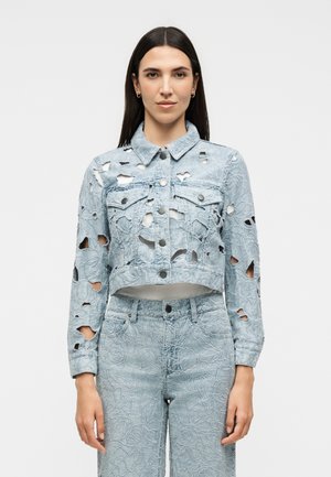 Vrouw draagt lichtblauwe geborduurde spijkerjas met grote gescheurde gaten en bijpassende hoge taille spijkerbroek tegen een effen achtergrond.