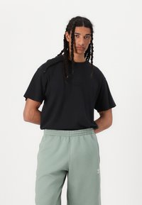 Μαύρο βαμβακερό t-shirt με κοντά μανίκια και ανάγλυφο λογότυπο, συνδυασμένο με ελαστικά παντελόνια σε ανοιχτό πράσινο χρώμα με μικρή λευκή λεπτομέρεια επωνυμίας.