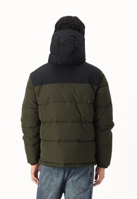 Puffet jakke med sort hætte og overdel, olivenfarvet krop, kviltet tekstur og elastiske manchetter, parret med blå denim jeans.