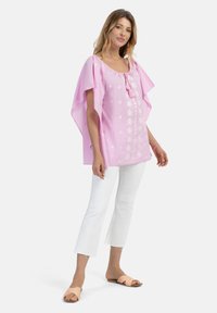 Lyserød bomulds-tunika med løs pasform, prydet med broderede blomsterdesigns og en bindesløjfe ved halsen. Båret med hvide ankelbukser og sandaler.