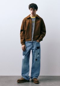 Bruine suède jas over een gestreept shirt, gecombineerd met lichtblauwe distressed jeans met patchwork, en bruine schoenen met een dik profiel.