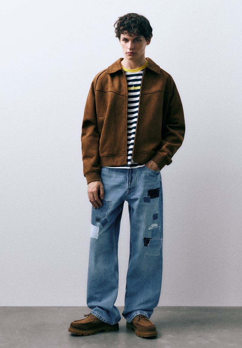 Bruine suède jas over een gestreept shirt, gecombineerd met lichtblauwe distressed jeans met patchwork, en bruine schoenen met een dik profiel.