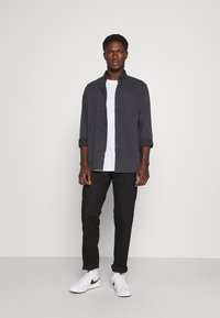 Camisa de botones gris oscuro sobre una camiseta blanca, combinada con jeans negros y zapatillas blancas. La camisa tiene un bolsillo de parche y mangas enrolladas.