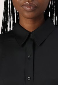Blusa preta com colarinho pontudo, frente abotoada e textura de tecido suave. Apresenta botões de plástico pretos e um design ajustado.
