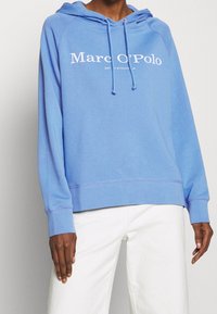 Felpa blu con cappuccio e logo bianco "Marc O'Polo" sul petto. Tessuto morbido, tasca a canguro e polsini a coste.