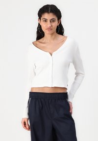 Weißer gerippter, kurzer Cardigan mit V-Ausschnitt, Knopfleiste und langen Ärmeln, kombiniert mit navyblauen Hose mit elastischem Bund.