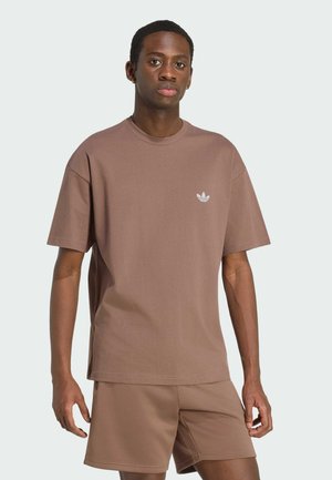 Ung mand iført brun Adidas t-shirt og matchende shorts, stående mod en ensfarvet lys baggrund.
