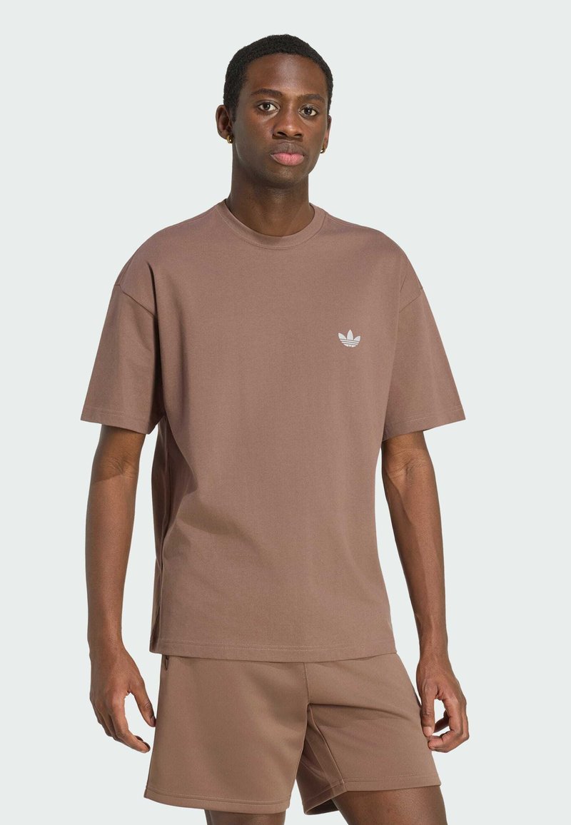 Jeune homme portant un t-shirt Adidas marron et un short assorti, debout devant un fond clair uni.