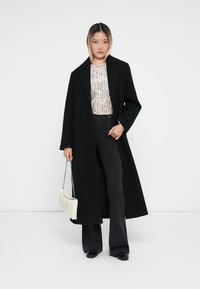 Abrigo largo negro, blusa de patrones claros, pantalones de campana negros, sosteniendo un bolso blanco con cadena. Texturas suaves, diseño a medida.