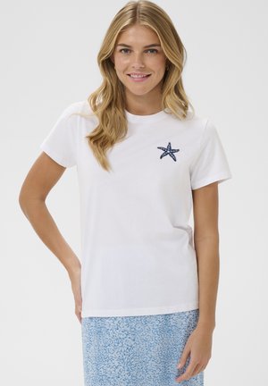 Smilende kvinde med blondt hår iført en hvid t-shirt med et lille blåt søstjernedesign og en blå mønstret nederdel, stående foran en ensfarvet baggrund.