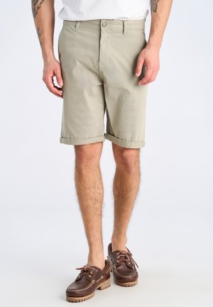 Mann trägt beige hochgekrempelte Shorts, weißes Hemd und braune Leder-Bootsschuhe, steht vor einem schlichten hellen Hintergrund.