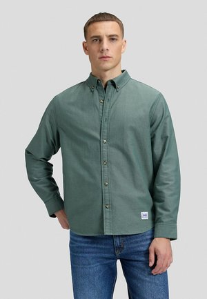 Uomo in piedi davanti a uno sfondo semplice, indossa una camicia verde a maniche lunghe con bottoni e jeans blu, rivolto in avanti con un'espressione neutra.