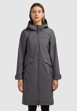 Frau trägt einen langen, dunkelgrauen Wintermantel mit Kapuze, Frontreißverschluss und Klappentaschen, steht vor einem einfarbigen hellgrauen Hintergrund.