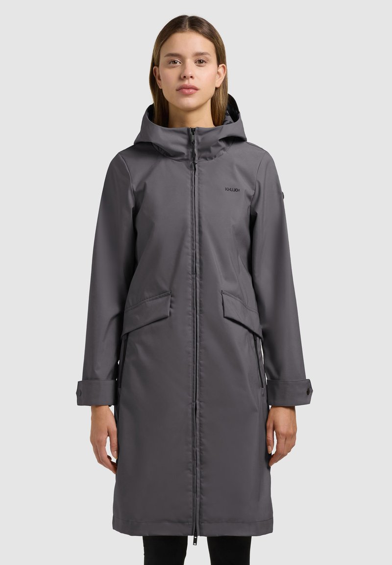 Mujer con un abrigo de invierno largo, gris oscuro, con capucha, cremallera frontal y bolsillos con solapa, posando frente a un fondo liso de color gris claro.