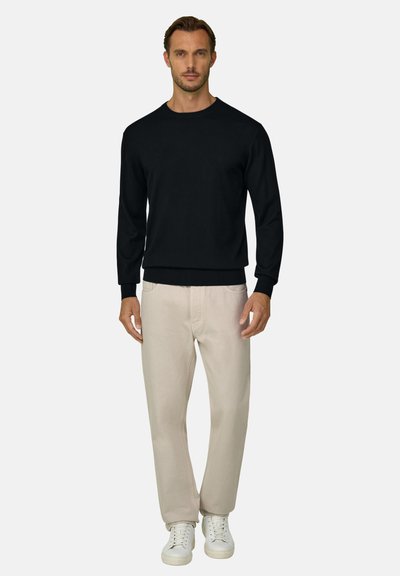 Homme debout portant une chemise noire à manches longues, un pantalon beige et des baskets blanches sur un fond uni gris clair.