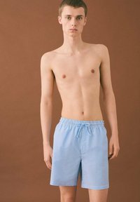 Modre in bele črtaste kopalne hlače z elastičnim pasom in podrobnostjo z vezalko. Moški model brez majice stoji pred rjavim ozadjem.