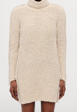 Beige textureret langærmet sweaterkjole med rullekrave, båret af en person med mørk hud, hænderne afslappede langs siderne.