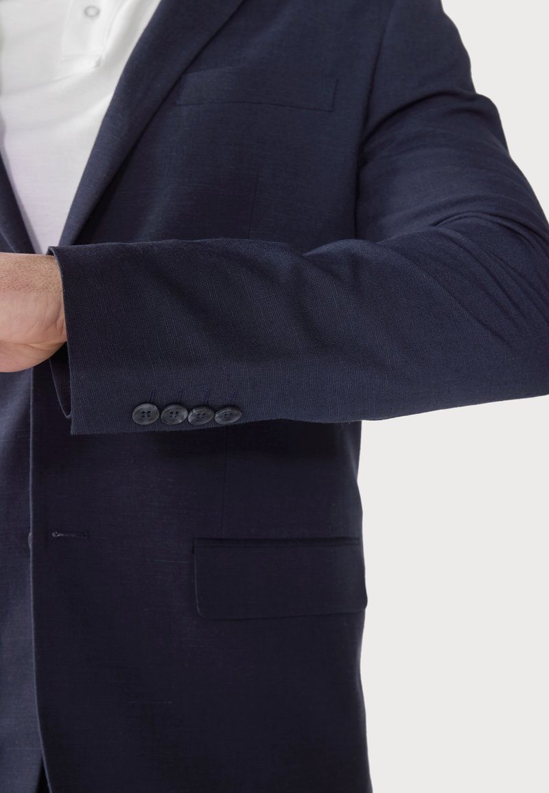 Blazer bleu marine en tissu texturé, avec des poignets à quatre boutons et une poche poitrine, accompagné d'une chemise blanche en dessous.