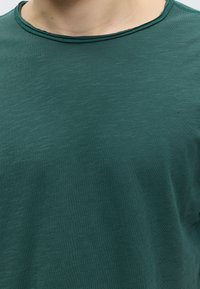 Κοντινή λήψη ενός ατόμου που φοράει πράσινο μπλουζάκι με στρογγυλή λαιμόκοψη, με ορατή υφή υφάσματος και ραφές γύρω από το λαιμό.